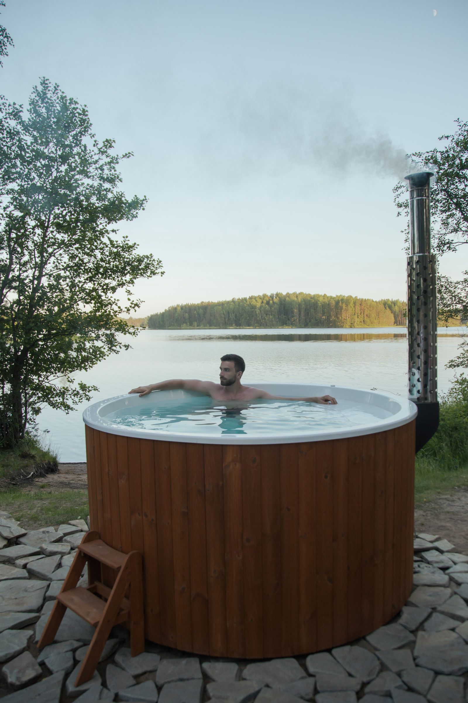 Man relaxing in a wooden hot tub beside a calm lake on a clear day